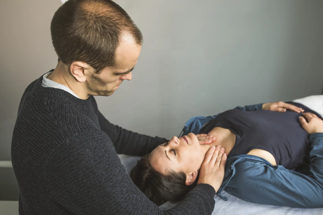 A person is receiving a therapeutic treatment while lying on a massage table, as another individual performs a technique at their head.