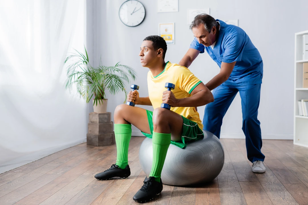 A person in a yellow sports shirt and green shorts performs squats on a stability ball while holding dumbbells, guided by a trainer.